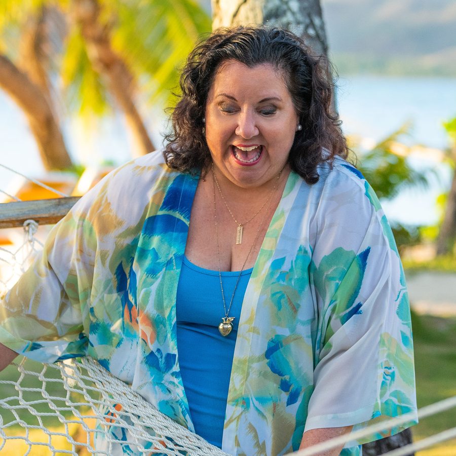 Dr. Lisa Hartwell laughing beside a hammock by the ocean in Hawaii, wearing her gold apple pendant